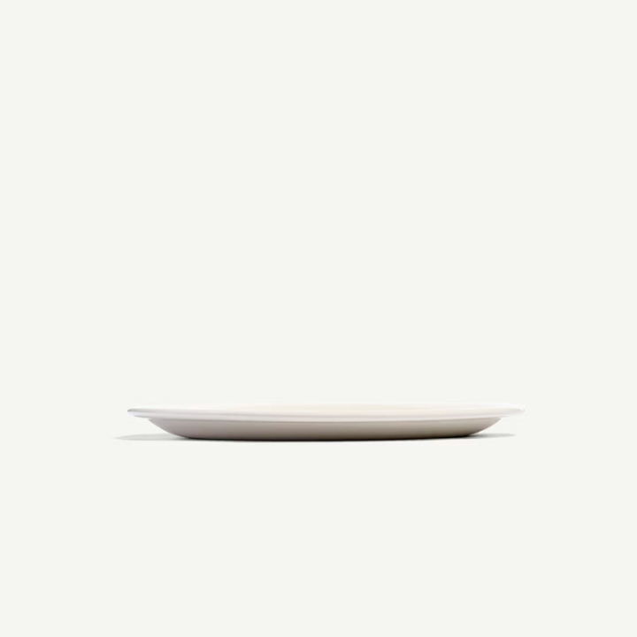 A minimalist, shallow oval plate in a matte off-white finish, photographed from a low angle against a solid, very light cream background.