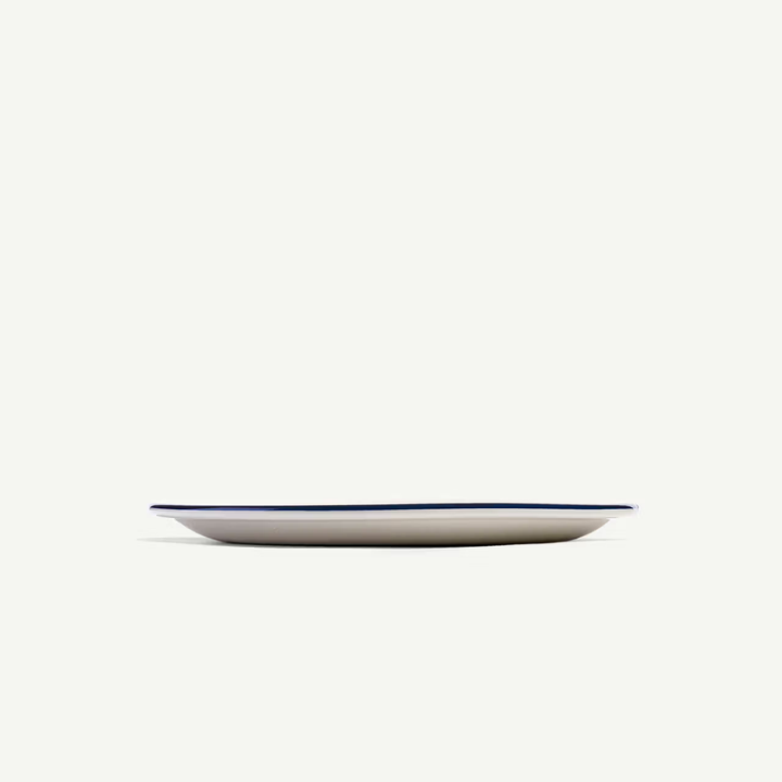 A minimalist oval ceramic plate with a thin dark blue rim, shown from a side profile against a solid off-white background.