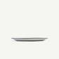 A minimalist oval ceramic plate with a thin dark blue rim, shown from a side profile against a solid off-white background.
