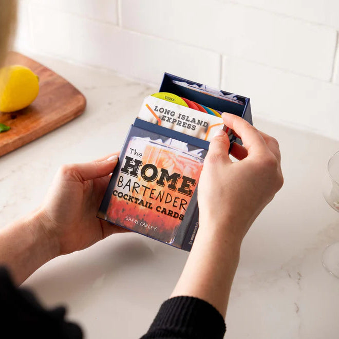Person holding 'The Home Bartender' cocktail card pack on a kitchen counter with a bottle and glass in the background.