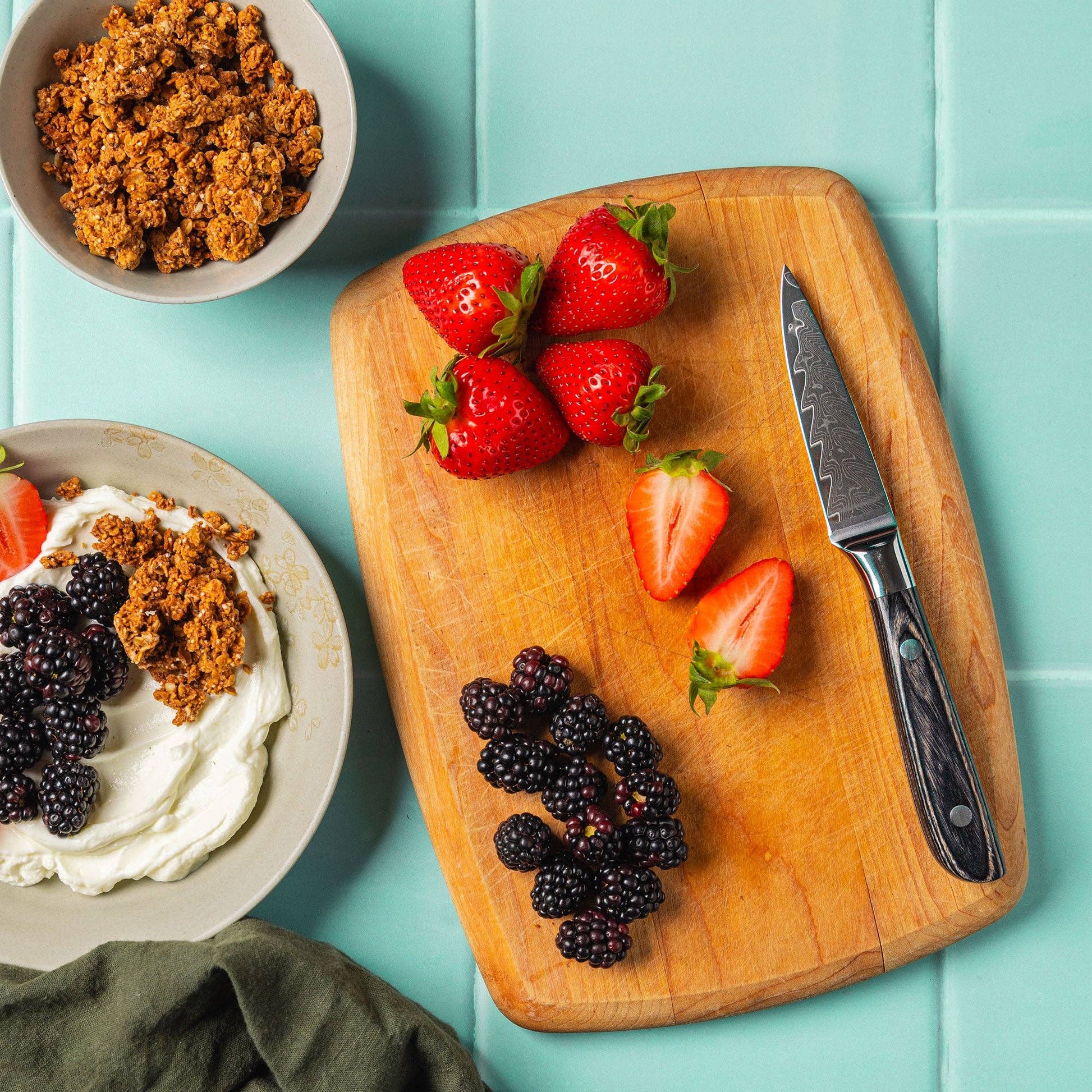 knife on cutting board with strawberries and black berries on a blue surface with bowls of yogurt and granola.