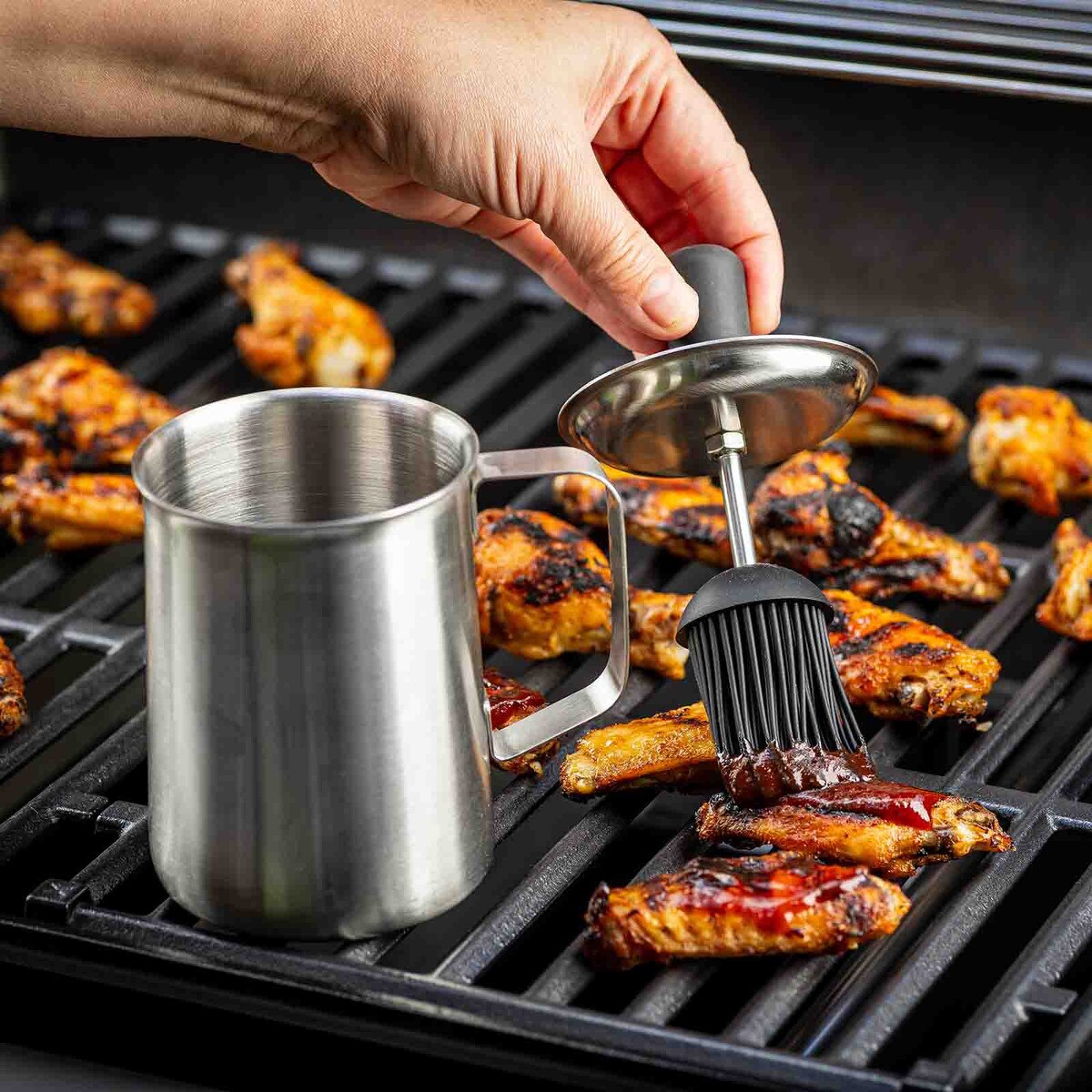 Person brushing barbecue sauce on grilled chicken wings with a metal brush and container.