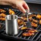 Person brushing barbecue sauce on grilled chicken wings with a metal brush and container.