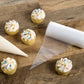 Decorated cupcakes with white frosting and colorful sprinkles on a wooden surface, with piping bags and paper in the foreground.