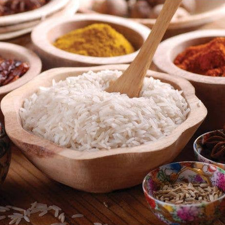 Wooden bowl of white rice with a wooden spoon, surrounded by spices on a wooden surface.