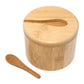 A round bamboo container with a matching lid featuring a cutout for a small wooden spoon, shown alongside a separate wooden spoon on a white background.