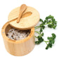 A round bamboo salt container open, showing coarse salt inside, with a small wooden spoon on the lid. Parsley sprigs are beside it on a white background.