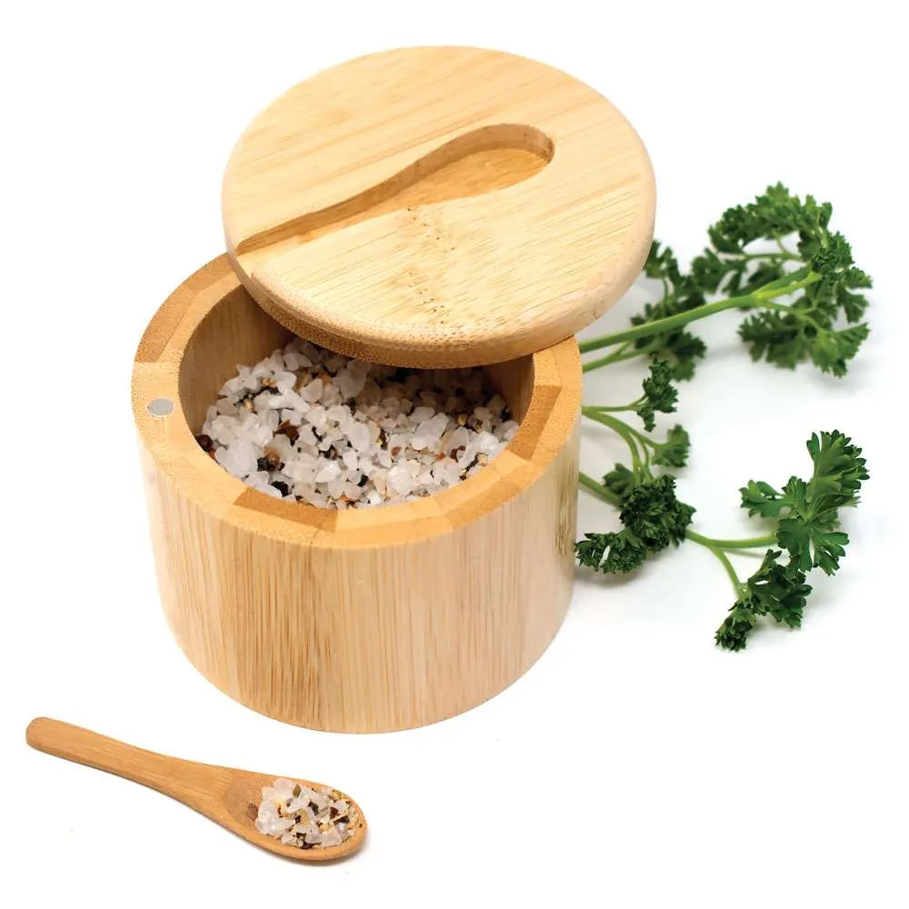 A round bamboo salt and pepper container with its lid off, filled with salt and pepper, on a white background with a small bamboo spoon and green parsley sprigs.