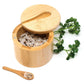 A round bamboo salt and pepper container with its lid off, filled with salt and pepper, on a white background with a small bamboo spoon and green parsley sprigs.