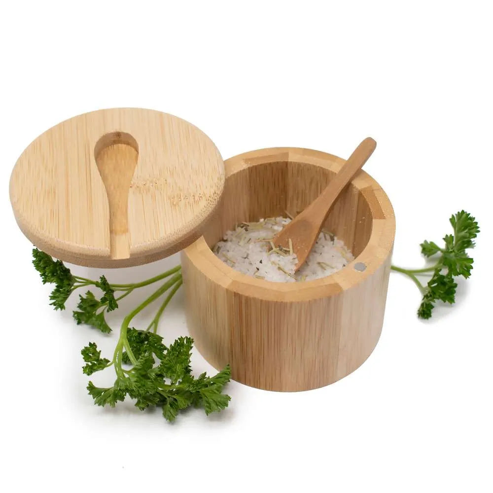 A bamboo salt and pepper container open, showing a small wooden spoon filled with white flakes. Fresh green parsley sprigs around the base on a white background.
