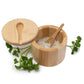 A bamboo salt and pepper container open, showing a small wooden spoon filled with white flakes. Fresh green parsley sprigs around the base on a white background.