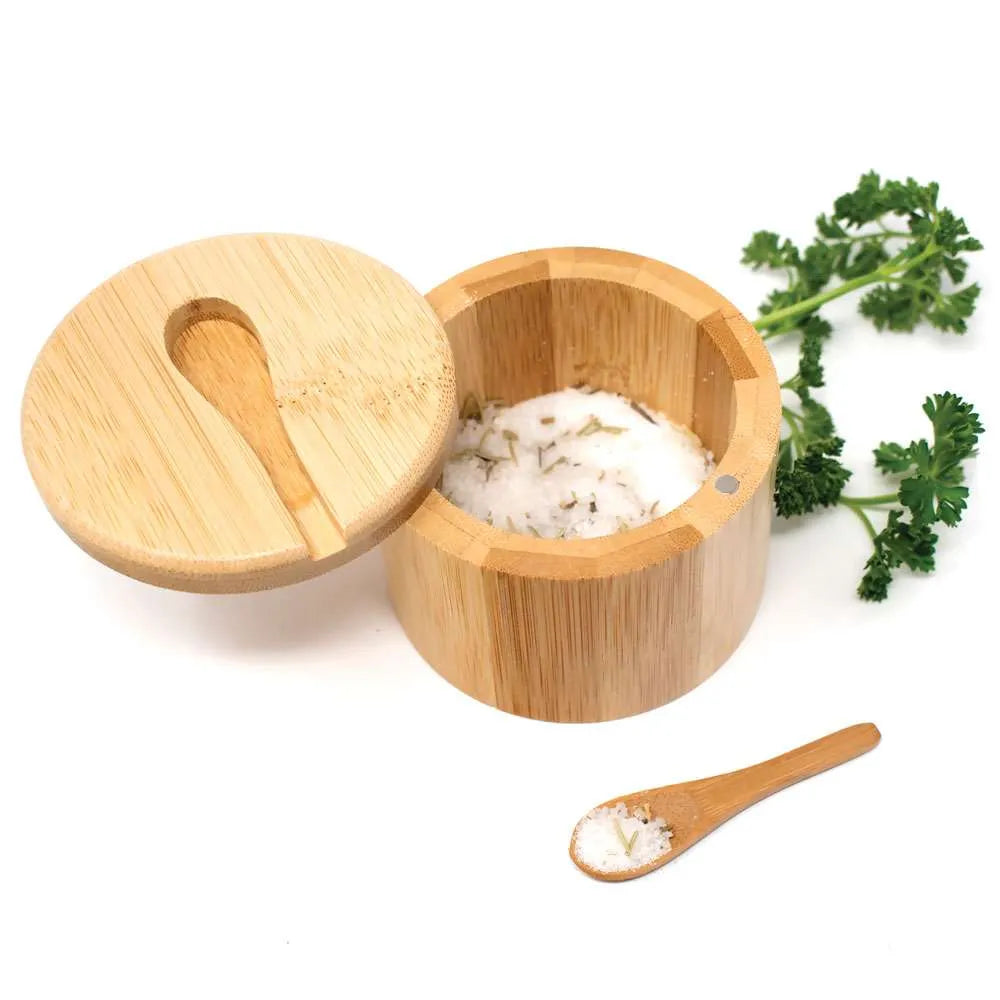 A round bamboo salt and pepper container open, showing white salt inside, and a small wooden spoon and fresh parsley on a white background.