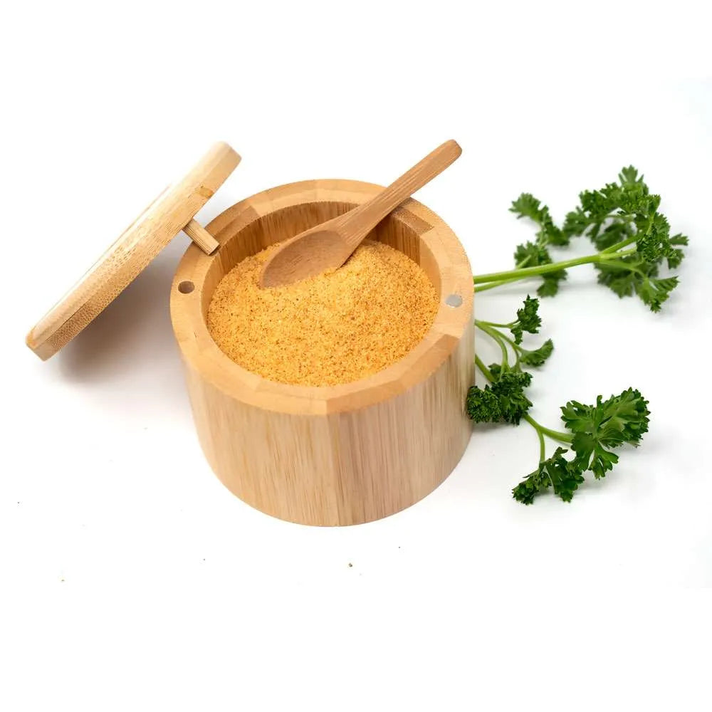 A light natural bamboo cylindrical spice container open, with yellow-orange ground spice and a small wooden spoon, with parsley on a white background.