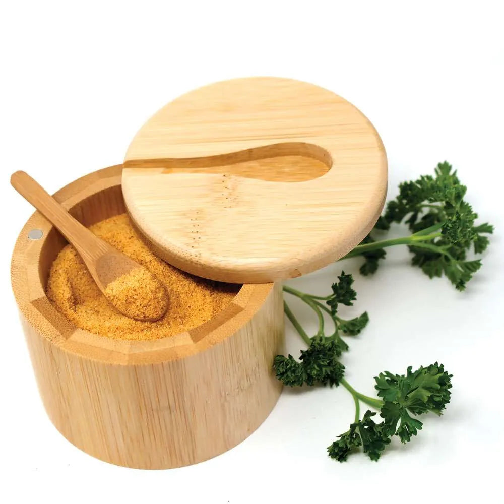 A round bamboo spice container with its lid propped open, showing a wooden spoon filled with yellow-orange ground spice. A small sprig of green parsley sits beside the container on a clean white background.