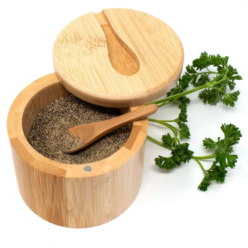 A round bamboo salt or pepper container with its lid propped open, showing dark ground seasoning inside. A small wooden spoon rests in the container, and fresh green parsley sprigs are placed beside it on a white background.