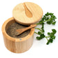 A round bamboo salt or pepper container with its lid propped open, showing dark ground seasoning inside. A small wooden spoon rests in the container, and fresh green parsley sprigs are placed beside it on a white background.