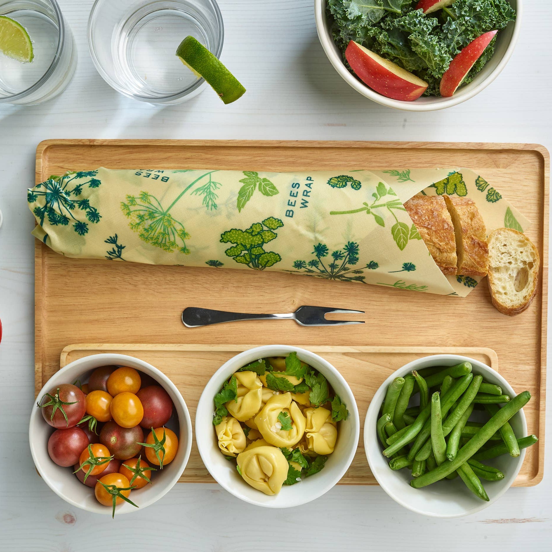 Wooden cutting board with green wrap, bowls of vegetables, and bread on a white surface.