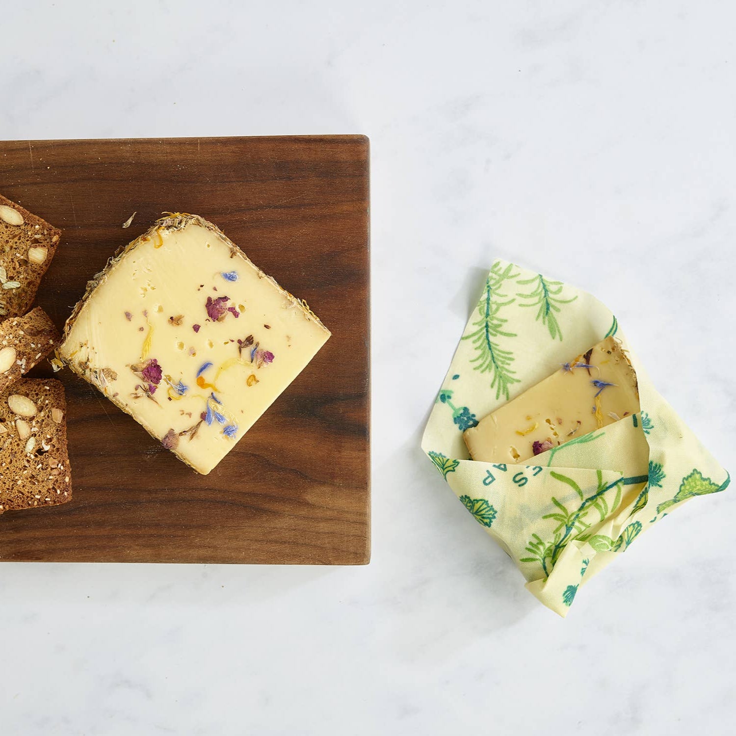 Wooden board with crackers and cheese, next to a reusable beeswax wrap on a white background