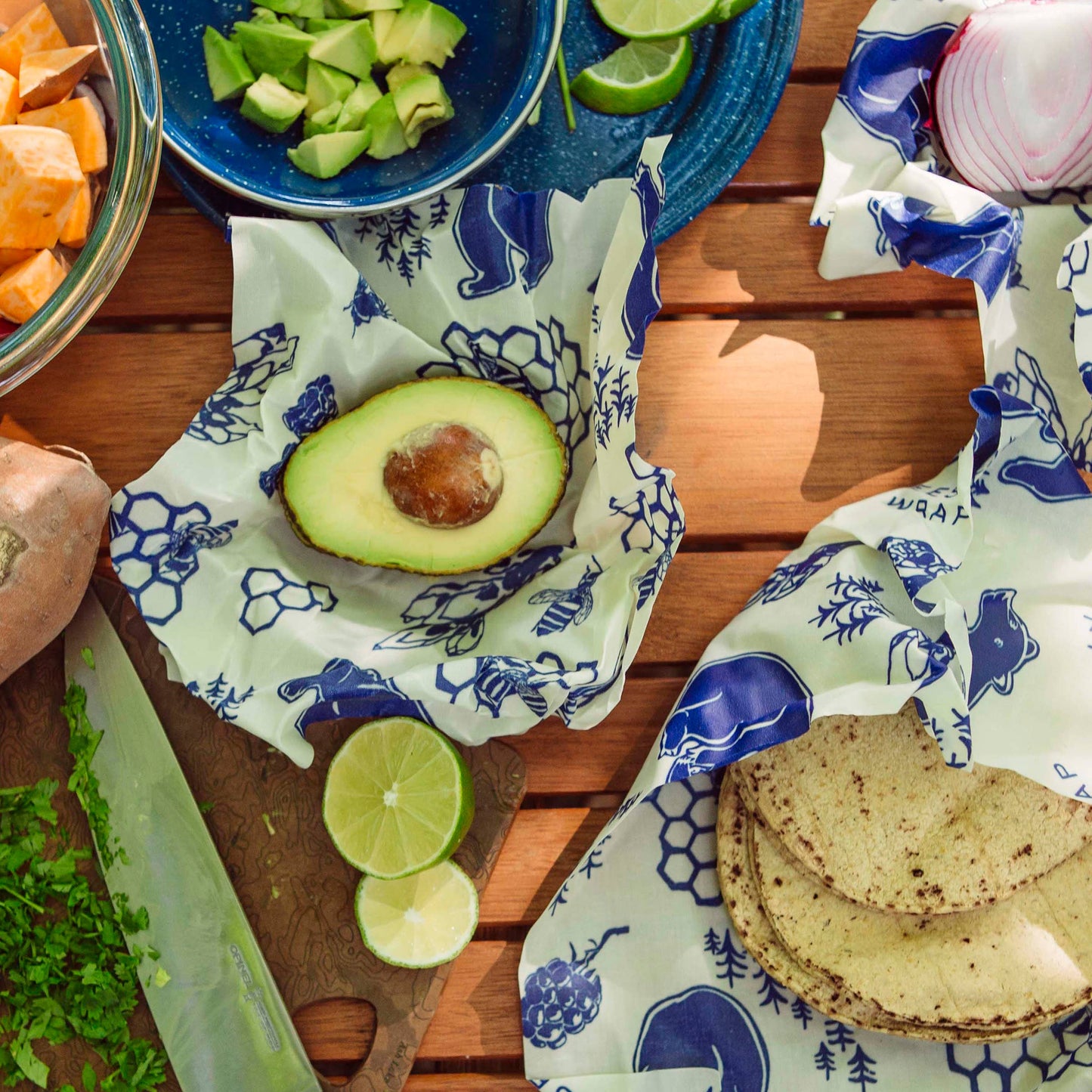 Avocado, tortillas, and lime halves on a wooden table with beeswax wraps.
