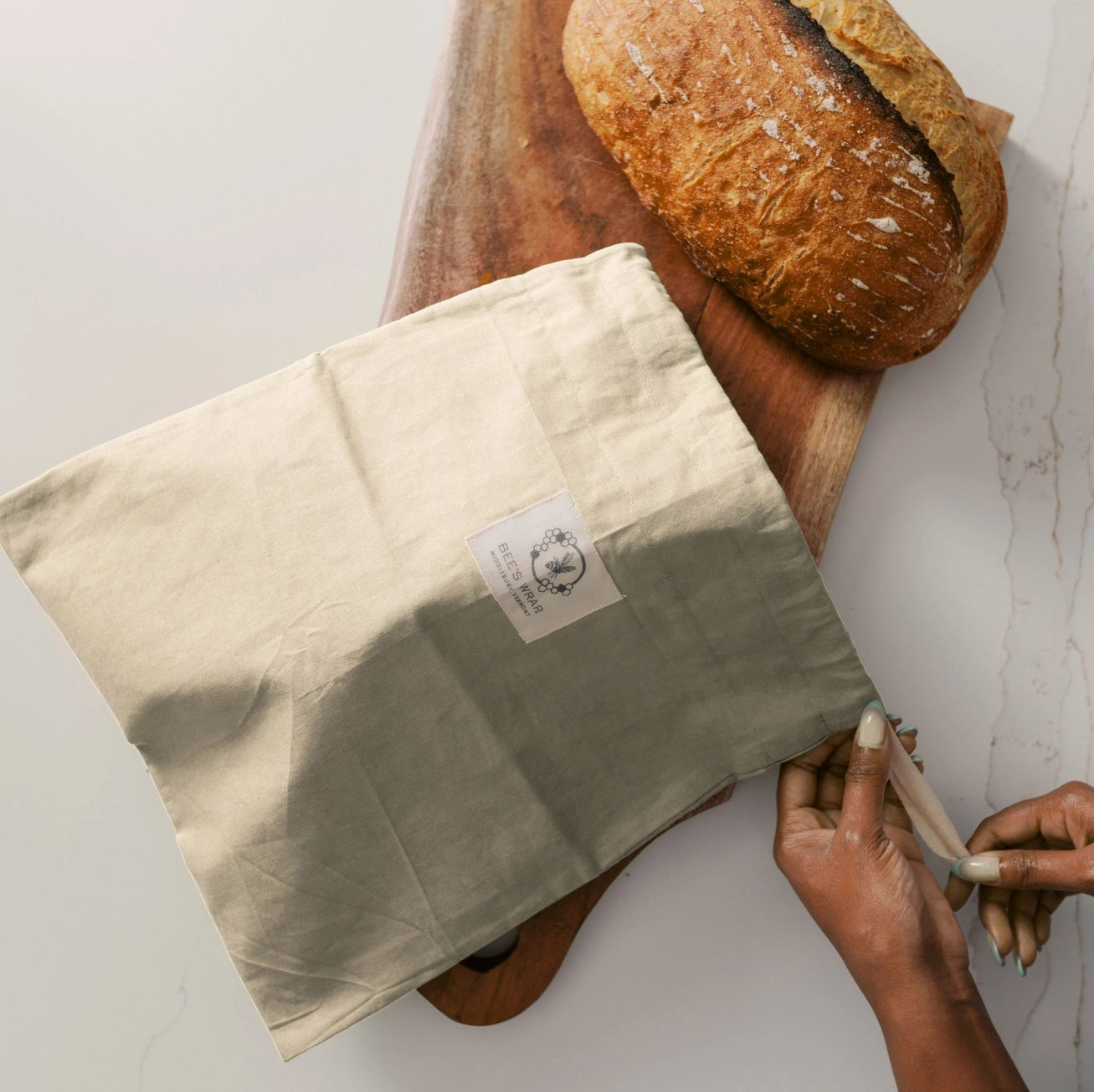 Person wrapping a loaf of bread in a cloth with a visible brand label on a white surface.