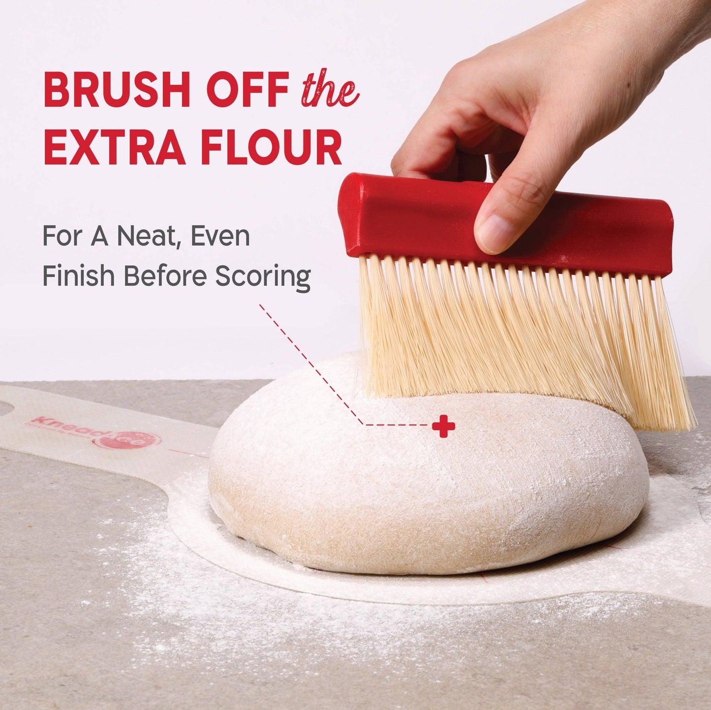Hand using a red brush to brush off extra flour from a dough ball on a light surface.