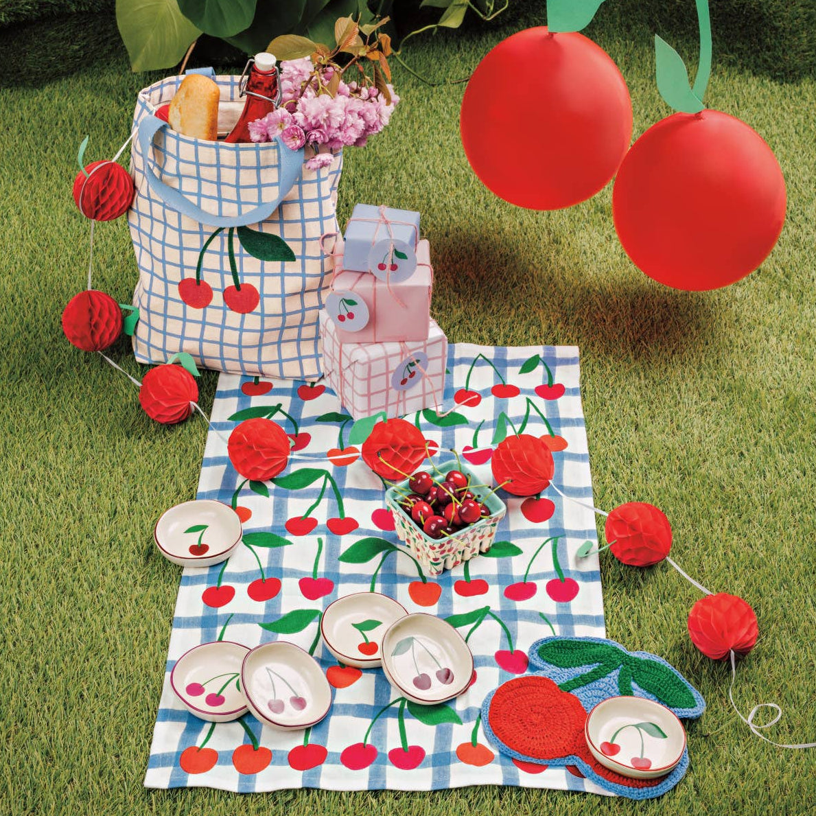Picnic setup with a checkered blanket, basket, and cherry-themed items on grass.