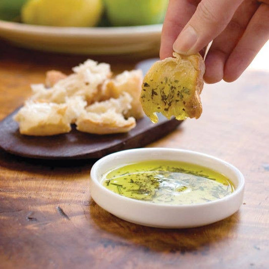 Person dipping a bread roll into a bowl of olive oil and herbs on a wooden table.