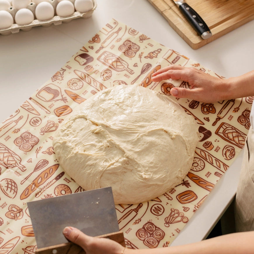 Bread dough on a patterned surface with hands working on it, surrounded by baking items.