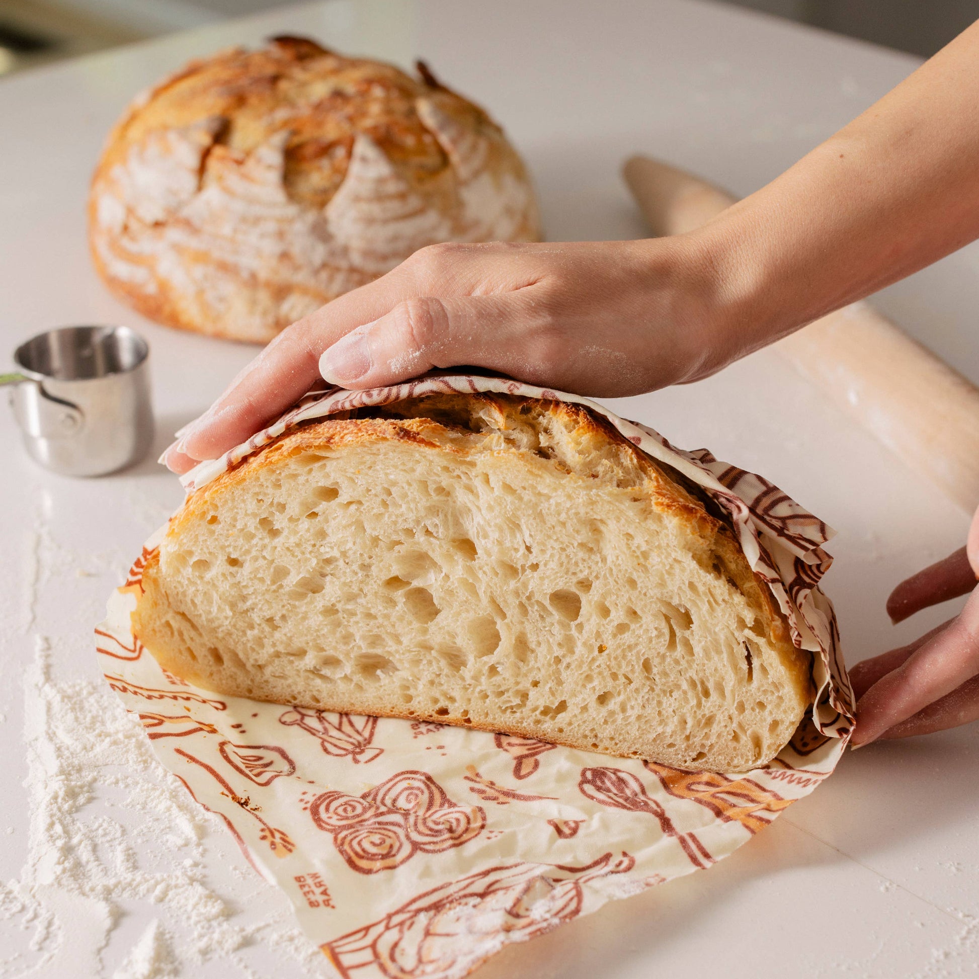 Person slicing a loaf of bread on a decorative cutting board with another loaf in the background.