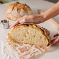 Person slicing a loaf of bread on a decorative cutting board with another loaf in the background.