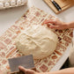 Bread dough on a patterned surface with hands working on it, surrounded by baking items.