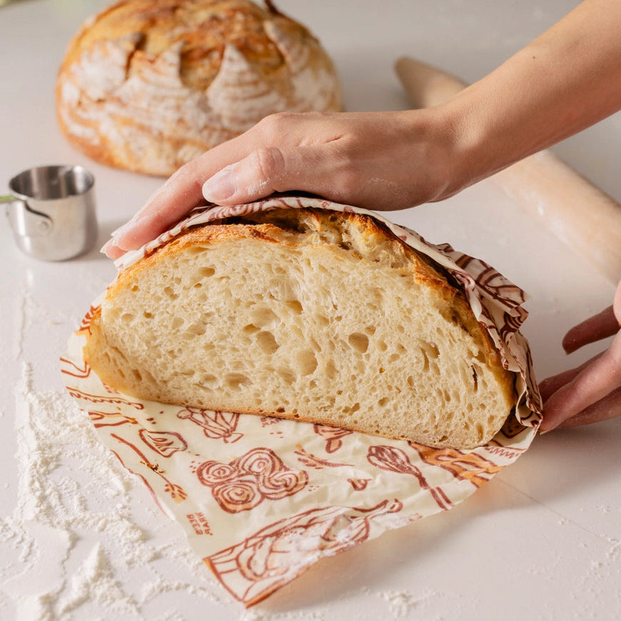 Person slicing a loaf of bread on a decorative cutting board with another loaf in the background.