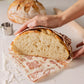 Person slicing a loaf of bread on a decorative cutting board with another loaf in the background.