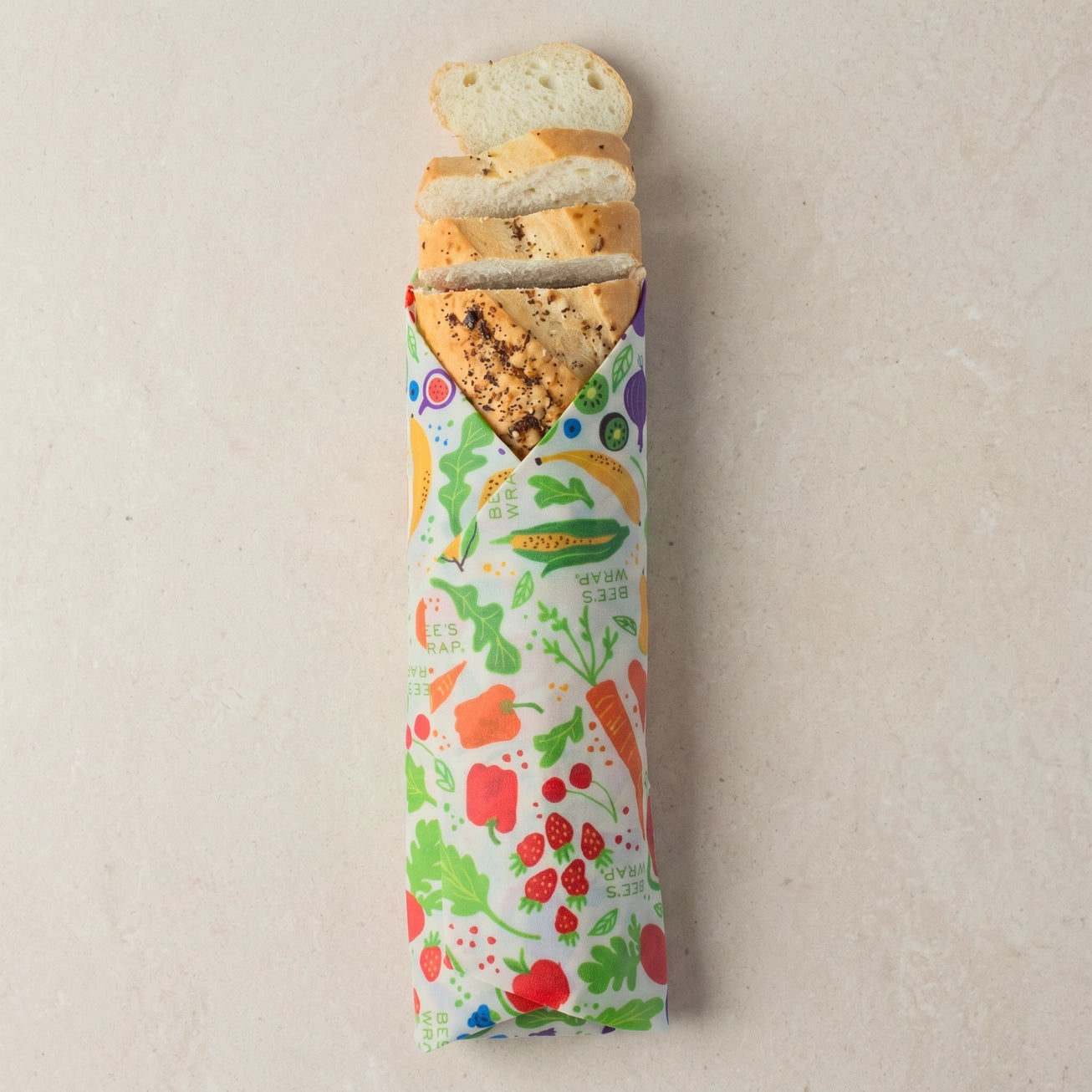 A loaf of bread wrapped in a colorful vegetable-print paper sleeve, with several slices cut and fanned out at the top, photographed on a light beige textured background.