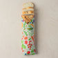 A loaf of bread wrapped in a colorful vegetable-print paper sleeve, with several slices cut and fanned out at the top, photographed on a light beige textured background.