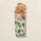 A loaf of rustic bread partially wrapped in a reusable fabric bread bag printed with botanical illustrations of herbs and leaves, set against a textured light beige background.