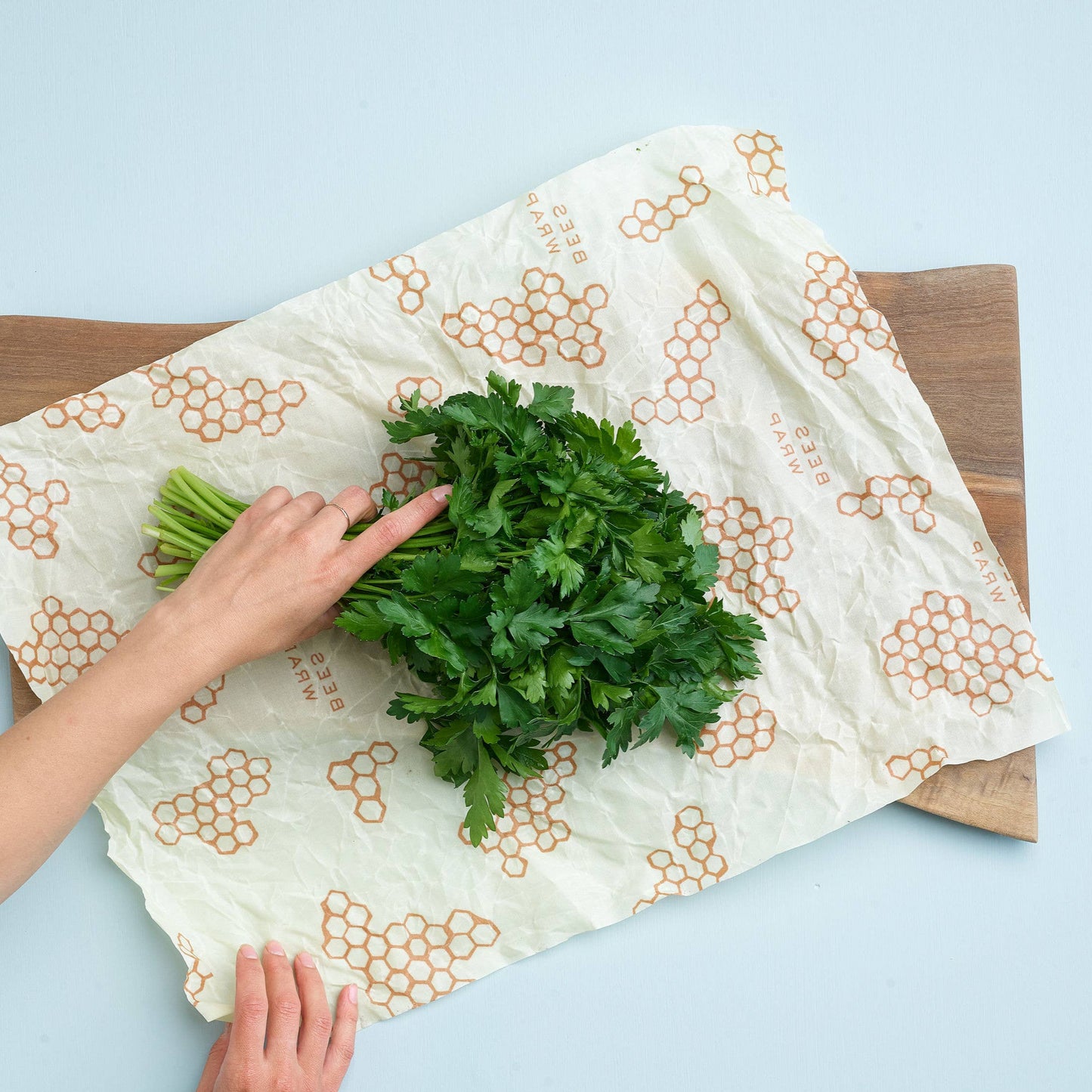 Person using a beeswax wrap to cover greens on a wooden cutting board with a light blue background