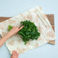 Person using a beeswax wrap to cover greens on a wooden cutting board with a light blue background