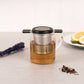 Glass teapot with metal infuser on a light surface with lavender and lemon slices.