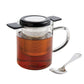 Clear glass teapot with black lid and handle, filled with tea, next to a silver spoon on a white background.