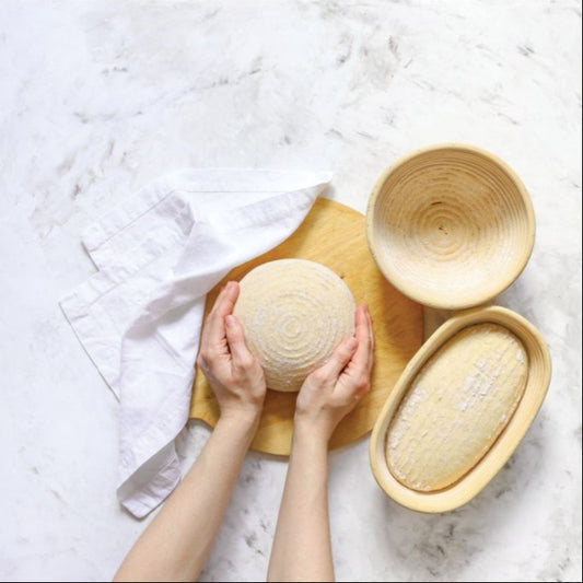 Hands holding a round bread loaf next to two wooden bread baskets on a white marble surface.