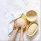 Hands holding a round bread loaf next to two wooden bread baskets on a white marble surface.