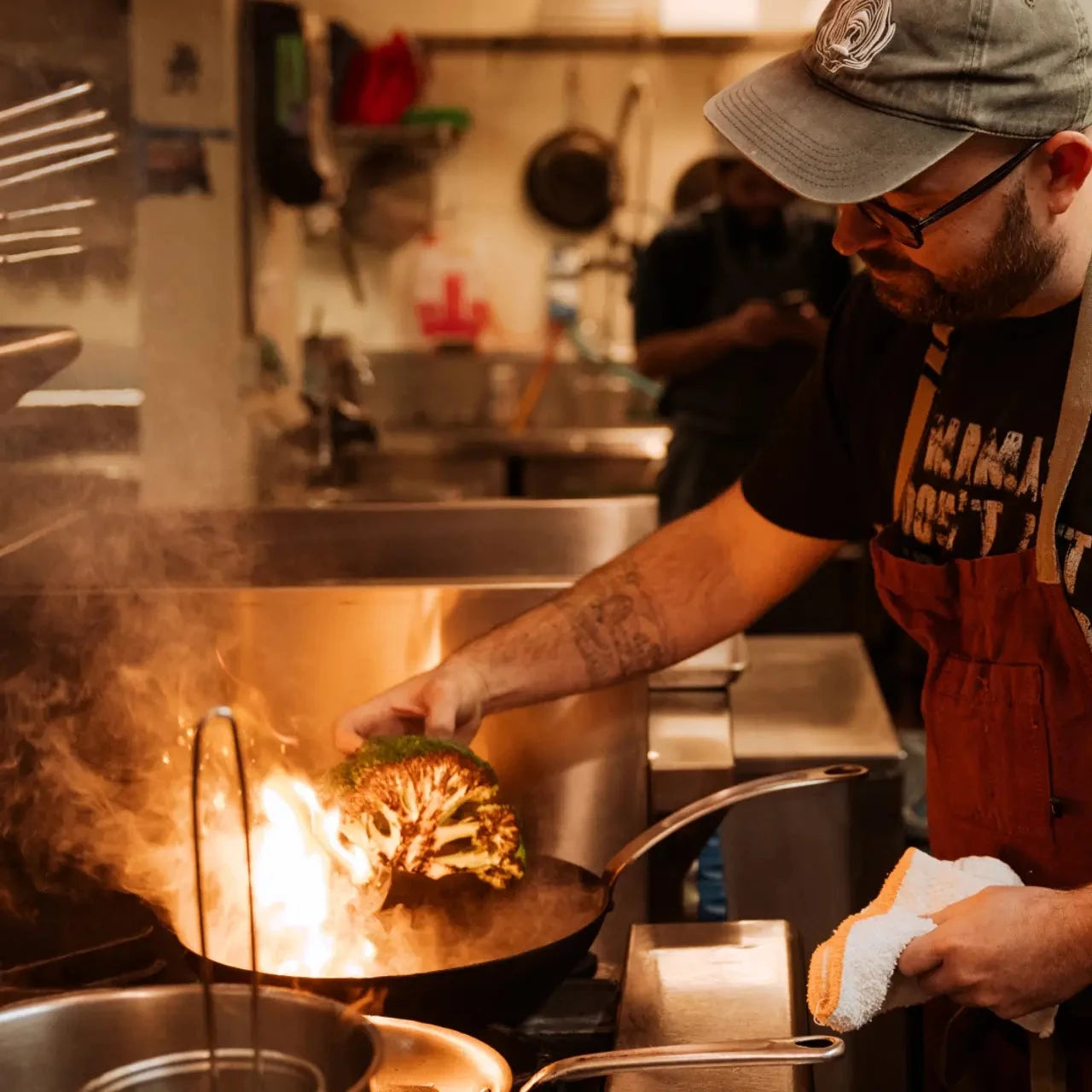 A black carbon steel frying pan being used to char a vegetable by a chef in a busy commercial kitchen.