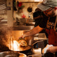 A black carbon steel frying pan being used to char a vegetable by a chef in a busy commercial kitchen.