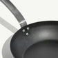 A black carbon steel frying pan with a stainless steel handle, pre-seasoned and ready for cooking on a white background.