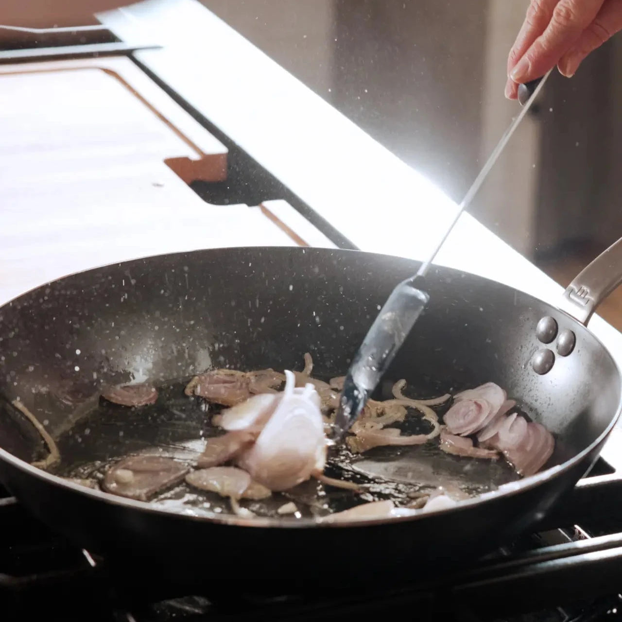 A black carbon steel frying pan with a stainless steel handle, with onions frying over a kitchen grill.