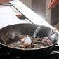 A black carbon steel frying pan with a stainless steel handle, with onions frying over a kitchen grill.