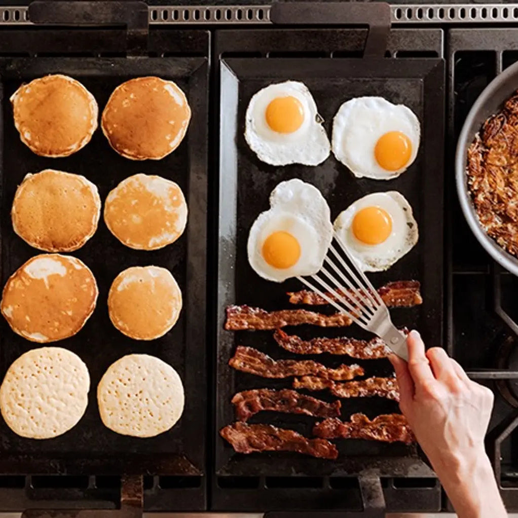 Cooking breakfast items on a griddle with pancakes, eggs, and bacon.