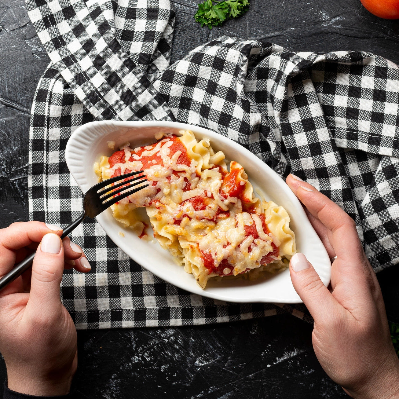A top-down view of a person's hands holding a white oval plate of pasta with tomato sauce and melted cheese, with one hand holding a black fork. The plate is placed on a black and white checkered cloth over a dark textured surface.