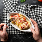 A top-down view of a person's hands holding a white oval plate of pasta with tomato sauce and melted cheese, with one hand holding a black fork. The plate is placed on a black and white checkered cloth over a dark textured surface.